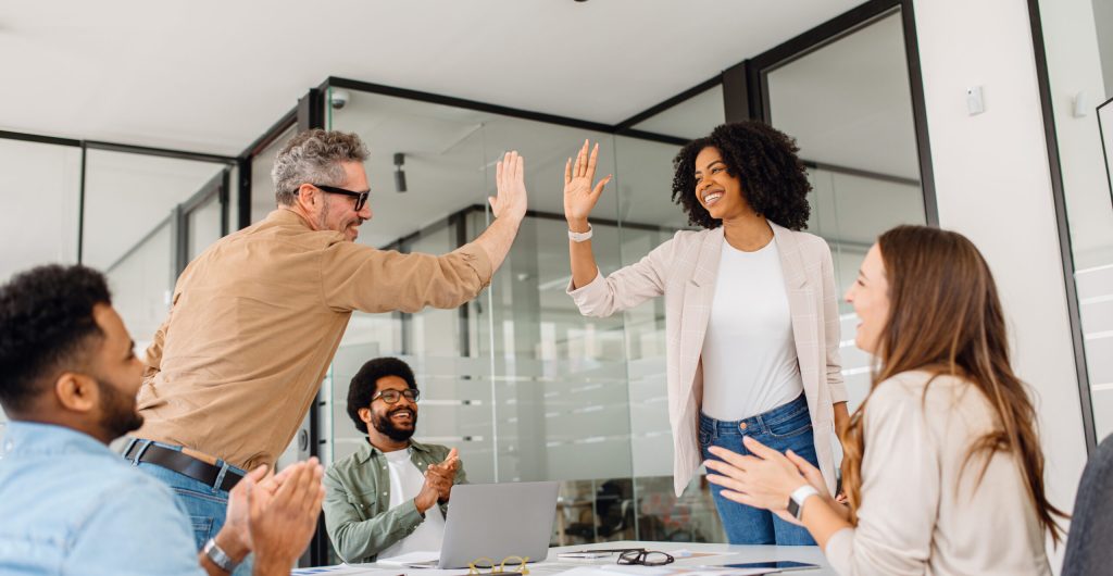 A diverse team of professionals celebrates a workplace achievement with a high-five in a modern office. The moment captures motivation, cooperation, and a positive business culture focused on teamwork