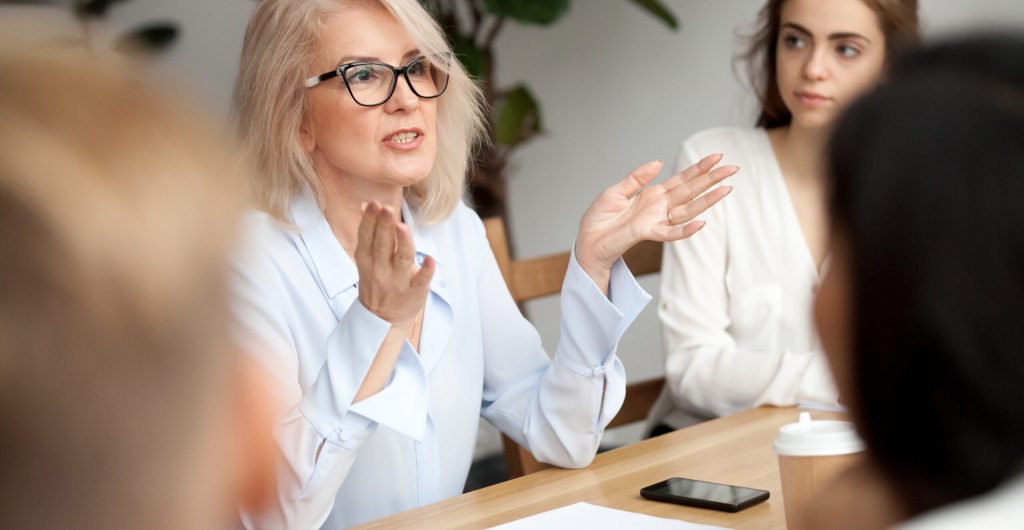 Attractive aged businesswoman, teacher or mentor coach speaking to young people, senior woman in glasses teaching audience at training seminar, female business leader speaker talking at meeting